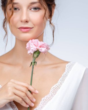 Açık tenli, doğal makyajlı, beyaz dantelli bir elbise giyerken elinde pembe bir karanfil tutan beyaz bir kadın. Güzellik, cilt bakımı, çiçek ve sağlık promosyonları için mükemmel..
