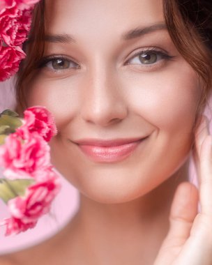 Beyaz tenli, zarif makyajlı ve pembe karanfilli beyaz bir kadının moda stüdyo güzelliği. Kozmetik endüstrisi görselleri, cilt reklamları ve modern güzellik markaları için mükemmel..