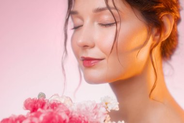 Kapalı gözlü, narin pembe ve beyaz bir çiçek buketinin kokusunun tadını çıkaran huzurlu genç bir kadının yakın plan portresi. Sıcak altın-pembe parıltı onun yumuşak özelliklerini güçlendirir ve bir...