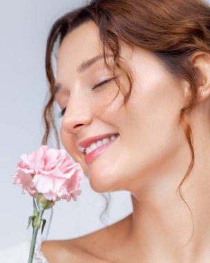 Beyaz dantelli bir elbise giyen zarif bir kadın gözleri kapalı gülümsüyor, elinde pembe bir karanfil tutuyor. Çiçek kokulu reklamlar, cilt bakımı promosyonları ve kadın yaşam tarzı görselleri için mükemmel..