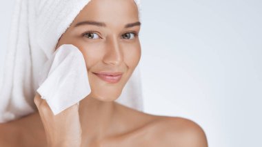 Saçına havlu sarılmış gülümseyen bir kadın nazikçe yüz silme kullanıyor. Taze bir cilt göstergesi. Güzellik, cilt bakımı ve sağlık ürünlerinin temizlenmesi ve temizlenmesi için ideal.