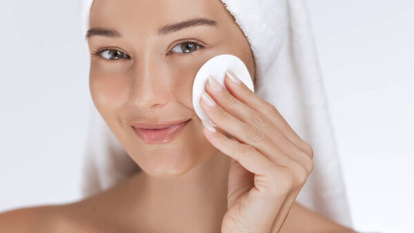 Улыбающаяся женщина нежно очищает лицо ватным блокнотом, демонстрируя свою здоровую, светящуюся кожу. Это изображение идеально подходит для кожи, красоты и велнес-контента, фокусируясь на уходе за лицом и сиянии.