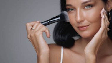 Gülümseyen bir kadının yakın plan portresi fırçayla makyaj yapıyor, cilt bakımını ve güzellik rutinini ön plana çıkarıyor. Güzellik markaları, makyaj dersleri ve kozmetik reklamları için ideal.