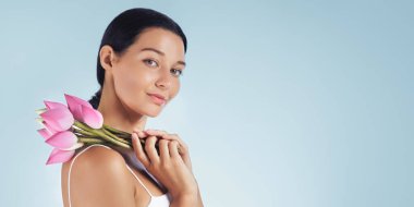 Zarif bir kadın omzunda pembe nilüfer tomurcuklarıyla poz veriyor, doğal güzelliği ve sağlığı vurguluyor. Spa tedavileri, cilt ürünleri reklamları ve lüks güzellik promosyonları için mükemmel.