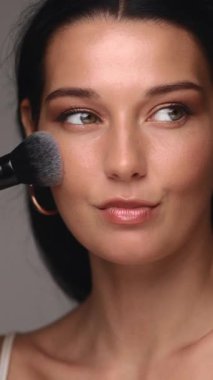 Koyu renk saçlı, tüylü fırça kullanarak makyaj yapan güzel bir kadının yakın plan fotoğrafı. Kusursuz bir teni, ince bir göz makyajı ve kendine güvenen bir ifadesi var. Güzellik ve kozmetik için mükemmel.