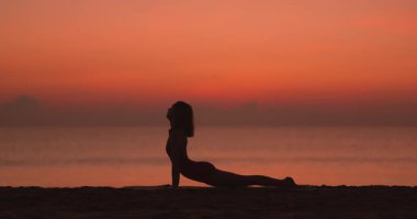Bir kadın gün batımında sakin bir plajda yoga pozları veriyor, siluette yakalanmış. Titrek gökyüzü ve sakin okyanus huzur dolu bir zemin oluşturur, sağlık, farkındalık ve zindelik temaları için mükemmel.. 