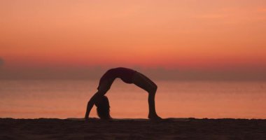 Bir kadın köprü pozu veriyor, bir bacağı kaldırılmış, silueti plajda gün batımına karşı parlıyor. Sakin okyanus ve canlı gökyüzü onun gücünü ve esnekliğini vurguluyor.