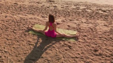 Bir kadın, güneş doğarken sakin okyanusa bakan kumlu bir plajda yoga pozunda tam bölünme pratiği yapıyor. Esneklik, farkındalık, açık hava fitness ve barışçıl sağlık inzivaları kavramları için mükemmel..