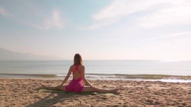 Sahilde oturmuş yoga yapan bir kadının arka görüntüsü. Gün doğumunda sakin okyanusa bakıyor. Rahatlama, farkındalık, sağIık ve sakin doğa ortamlarında açık hava fitness temaları için idealdir..