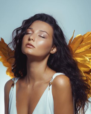 Şehvetli genç esmer kadın altın melek kanatları takıyor ve gözleri kapalı beyaz elbiseler giyiyor. Moda, güzellik veya kavramsal editörlük için mükemmel kaliteli stüdyo portresi.
