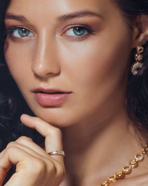 Zarif altın takılar takan ve göz alıcı makyaj yapan şık bir kadının yakın çekim stüdyo portresi. Kozmetik reklamlar, lüks promosyonlar ve modern güzellik kampanyaları için ideal..