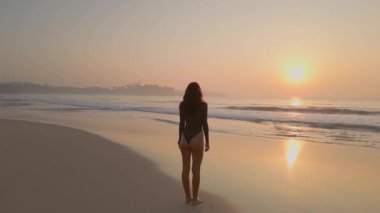 Mayo giymiş bir kadın gün doğumunda tropik bir plajın kenarında sessizce durur ve okyanusu seyreder. Huzur ve netlik temalı farkındalık, duygu ve yaşam tarzı görüntüleri için idealdir..