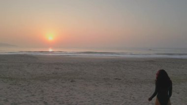 Güneşin doğuşuyla kumsalda yürüyen bronz tenli bir kadın, sakin bir okyanus manzaralı. Seyahat ve yaşam tarzı görselleri, plaj temalı projeler ve yüksek kaliteli kavramsal hikaye anlatımı için mükemmel..