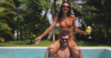 Tropikal bir yüzme havuzunun keyfini çıkaran oyuncu bir çift. Turuncu bikinili bir kadın içki tutarken erkeğin omuzlarında oturuyor, gülüyor ve kollarını açıyor. Yaz, tatil ve yaşam tarzı temaları için mükemmel. 