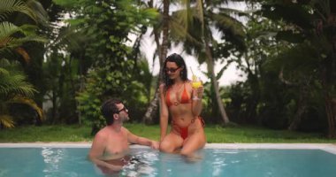 Bir çift, tropikal bir havuzda, turuncu bikinili kadın suyun içinde dururken elinde kokteyl tutarken rahatlatıcı bir sohbetin tadını çıkarıyor. Etrafı bereketli palmiye ağaçları ile çevrili, mükemmel bir tatil.