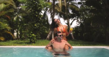 Tropik bir havuzda romantik bir çift sahnesi. Turuncu bikinili kadın yemyeşil bir ortamda erkeğin omuzlarında otururken kokteylden zevk alıyor. Tatil temaları için mükemmel. 