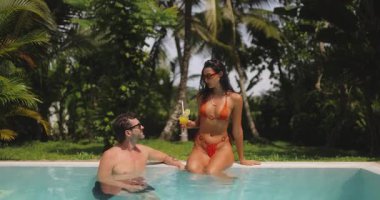 Bir çift, havuz başında turuncu bikinili bir kadının, sudaki adamla sohbet ederken elinde kokteyl tutmasıyla tropikal bir anın tadını çıkarıyor. Bereketli yeşillik ve palmiye ağaçları rahatlatıcı, tatili artırır