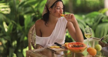 Şık yaz kıyafetleri içinde şık, bronzlaşmış bir kadın, tropik meyveler ve yemyeşil ile çevrili açık hava masasında bir bardak beyaz şarapla bir dilim portakalın tadını çıkarıyor. Yaşam tarzı temaları için mükemmel. 