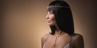 Pürüzsüz siyah saçlı, Kleopatra 'dan esinlenilmiş altın başlık ve kolye takan sofistike bir kadın, sıcak bir arka plana karşı zarif bir şekilde poz veriyor. Özgüvenli ifadesi ve lüks tarzı.