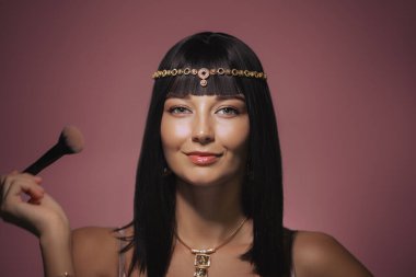 Parlak siyah saçlı, Kleopatra 'dan esinlenilmiş altın başlık ve kolye takan güzel bir kadın pembe arka planda makyaj fırçasını tutarken kendinden emin bir şekilde gülümsüyor. Onun ışıl ışıl görünüşü ve