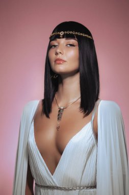 Kleopatra 'dan esinlenilmiş altın başlık ve kolye takan parlak siyah saçlı güzel bir kadın, pembe bir fona karşı güven içinde poz veriyor. Lüks tarzı ve özgüvenli ifadesi...