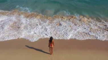 Bikinili bir kadının kumlu bir sahilde güçlü okyanus dalgalarıyla yüzleştiği destansı bir fotoğraf. Seyahat, macera ve tropikal yaşam tarzı kavramları için ideal. 