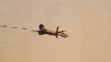 Arka planda turkuaz okyanus dalgaları olan kumlu bir plajda rahatlayan bikinili bir kadının hava görüntüsü. Tropikal tatil, yaz ve yaşam tarzı görüntüleri için mükemmel.. 