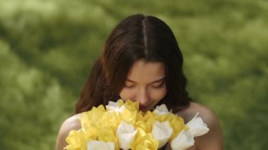 Uzun kahverengi saçlı, gülümseyen ve açık havada yemyeşil laleler gibi kokan güzel bir genç kadın. Doğa, güzellik, rahatlama ve refah temaları için kavramsal video.