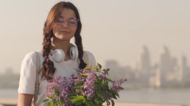 Gözlüklü, kulaklıklı ve leylak desenli genç bir kadın nehir kenarında huzurlu bir ilkbaharın tadını çıkarıyor. Yaşam tarzı, şehir doğası ve kadınsı sabah temaları için mükemmel..