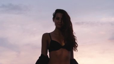Siyah bikinili ve deri ceketli çarpıcı genç bir kadın çarpıcı bir günbatımı gökyüzüne karşı güven içinde poz veriyor. Güçlendirme, moda ve doğal güzellik temaları için mükemmel.. 