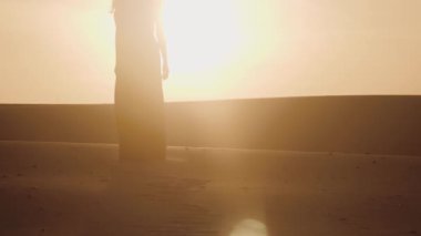 Çöl günbatımının altın parıltısına karşı dalgalı bir elbise giymiş nefes kesici bir kadın silueti. Sahne, yumuşak ışıkta güzellik, huzur ve sonsuz zarafeti yakalar..