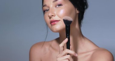 Parlak tenli kendine güvenen genç bir kadın elmacık kemiğine makyaj fırçası dayayıp doğal güzelliğini ve zarafetini vurguluyor. Güzellik, cilt bakımı ya da kozmetik reklamları için mükemmel.