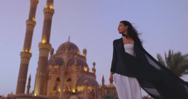 Şık giyinmiş zarif bir kadın, Mısır 'ın Sharm El Sheikh kentindeki güzel aydınlatılmış Sahaba Camii önünde alacakaranlıkta poz veriyor. Yüksek kaliteli kültürel ve seyahat promosyon içeriği için ideal.