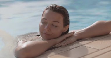 Bikinili bir kadın, gözleri kapalı ve gülümsüyor, havuz kenarında ağır çekimde ürperiyor. Yaşam tarzı, seyahat ve sağlık promosyonları için ideal, rahatlama, güzellik ve sükunetin vurgulanması.