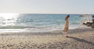 Türkiye 'nin Kemer kentindeki kayalık bir sahilde turkuaz dalgalar ve güneşli gökyüzüyle yürüyen zarif bir kadın. Ağır çekimde çekildi, seyahat promosyonları, yaşam tarzı ve rahatlama temaları için mükemmel.. 