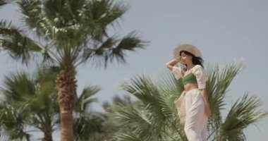 Palmiye ağaçları arasında tropikal tatil köyünde yaz kıyafetiyle yürüyen bir sahil kadını. Yüksek kalite ağır çekim 4k görüntü