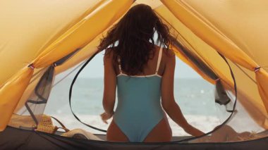 Mavi mayo giymiş bir kadın çadırın içinde ayağa kalkıp denize doğru yürürken arkadan görülüyor. Bu yüksek kaliteli görüntüler huzurlu bir sahil sahnesini çekiyor. Seyahat ve yaşam tarzı için mükemmel.