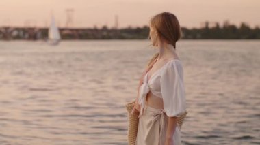 Beyaz bir elbise giymiş zarif bir kadın ve gün batımında nehir kenarında duran örgü bir çanta taşıyor. Yaz yaşam tarzı, seyahat kampanyaları ve romantik moda promosyonları için ideal.. 