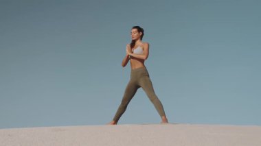 Genç bir kadın sakin çölde yoga yapıyor, kum tepelerinde duruyor ve açık mavi bir gökyüzünün altında zarafetle esniyor. Sağlık, zindelik ve meditasyon kavramları için ideal.. 