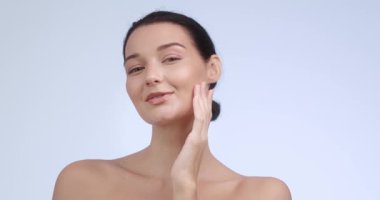 Gülümseyen beyaz kadın model, minimalist stüdyo ortamında nazikçe yanağını okşar. Lüks cilt bakımı, doğal güzellik ve kozmetik yüz bakımı için harika..