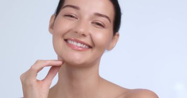Modayı takip eden genç bir kadın, minimalist stüdyo ışıklandırmasına tenini sürerken gülümser ve yüzüne dokunur. Etkilenen güzellik içeriği ve sosyal medya ürünü tanıtımı için ideal.