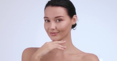 Beyaz bir kadın temiz bir stüdyoda yüzünü okşarken nazikçe gülümser. Dermatoloji reklamları, yaşlanma karşıtı cilt bakımı, yüz yenileme hizmetleri ve klinik güzellik kampanyaları için ideal..