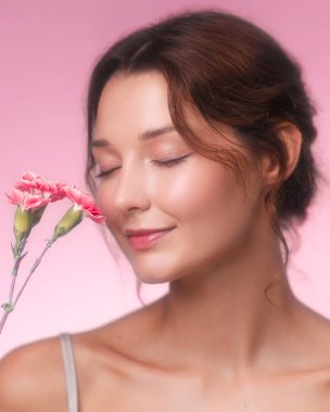 Yakın çekim stüdyo portresi, narin pembe çiçekleri koklarken nazikçe gülümseyen güzel bir genç kadın. Işıl ışıl cildi ve yumuşak makyajı pastel pembe arka planda parlıyor.