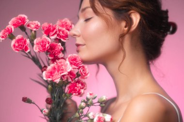 Kusursuz derisi ve kapalı gözleri olan zarif beyaz bir kadın pembe bir arka planda pembe karanfillerin tadını çıkarıyor. Cilt bakımı, kaplıca, çiçek güzelliği ve kişisel bakım temalı promosyonlar için ideal..
