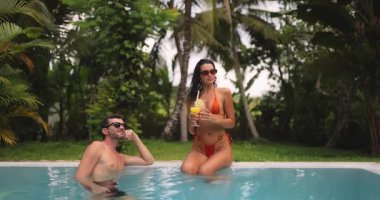 Havuz başında tropikal bir anın tadını çıkaran şık bir çift. Turuncu bikinili kadın, kenarda oturmuş, suda dinlenen bir adamla kokteyl tutuyor. Tatil temaları için ideal. 