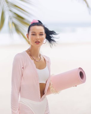 Yoga minderi taşıyan, tropikal bir plajda pembe bir giysi giyen modacı kadın. Spor, sağlık ve yaşam tarzı temaları için ideal, rahatlama ve dengeyi teşvik etmek