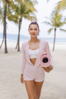 Pembe elbiseli kendine güvenen bir kadın tropik bir plajda yürürken yoga minderi tutuyor. Gülümsüyor ve günlük spor kıyafetleri giyiyor. Rahatlatıcı bir açık hava spor seansı öneriyor. Tam oldu.