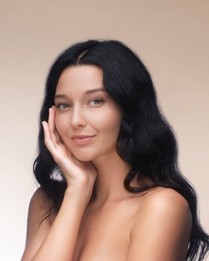 Uzun siyah saçlı dingin bir genç kadın, kusursuz cildini ve doğal güzelliğini göstermek için elini nazikçe yüzüne koyuyor. Bu resim cilt bakımı, sağlık ve güzellik ürünleri için idealdir.