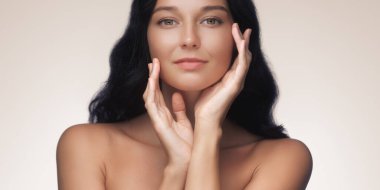 Uzun siyah saçlı zarif bir Asyalı kadın elleri yüzünün yanında zarifçe poz veriyor, kusursuz bir ten ve doğal güzellik sergiliyor. Güzellik, cilt bakımı ve kozmetik reklamları için ideal.