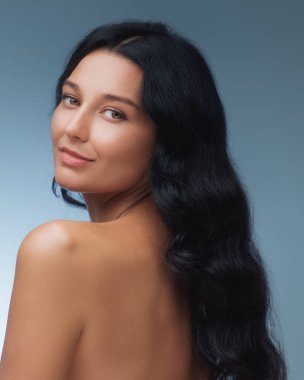 Uzun siyah saçlı çarpıcı bir kadın, yumuşak mavi bir arka planda pürüzsüz cildini ve doğal güzelliğini ön plana çıkarıyor. Güzellik, cilt bakımı ve sağlık konuları için ideal.. 